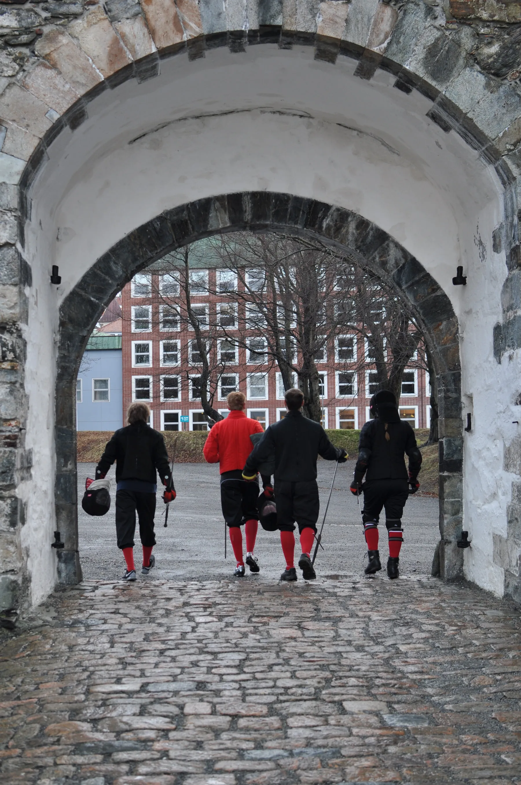 Fire fektere med sverd over skulderen går gjennom en steinhvelving på Bergenhus, med Bryggen i bakgrunnen.